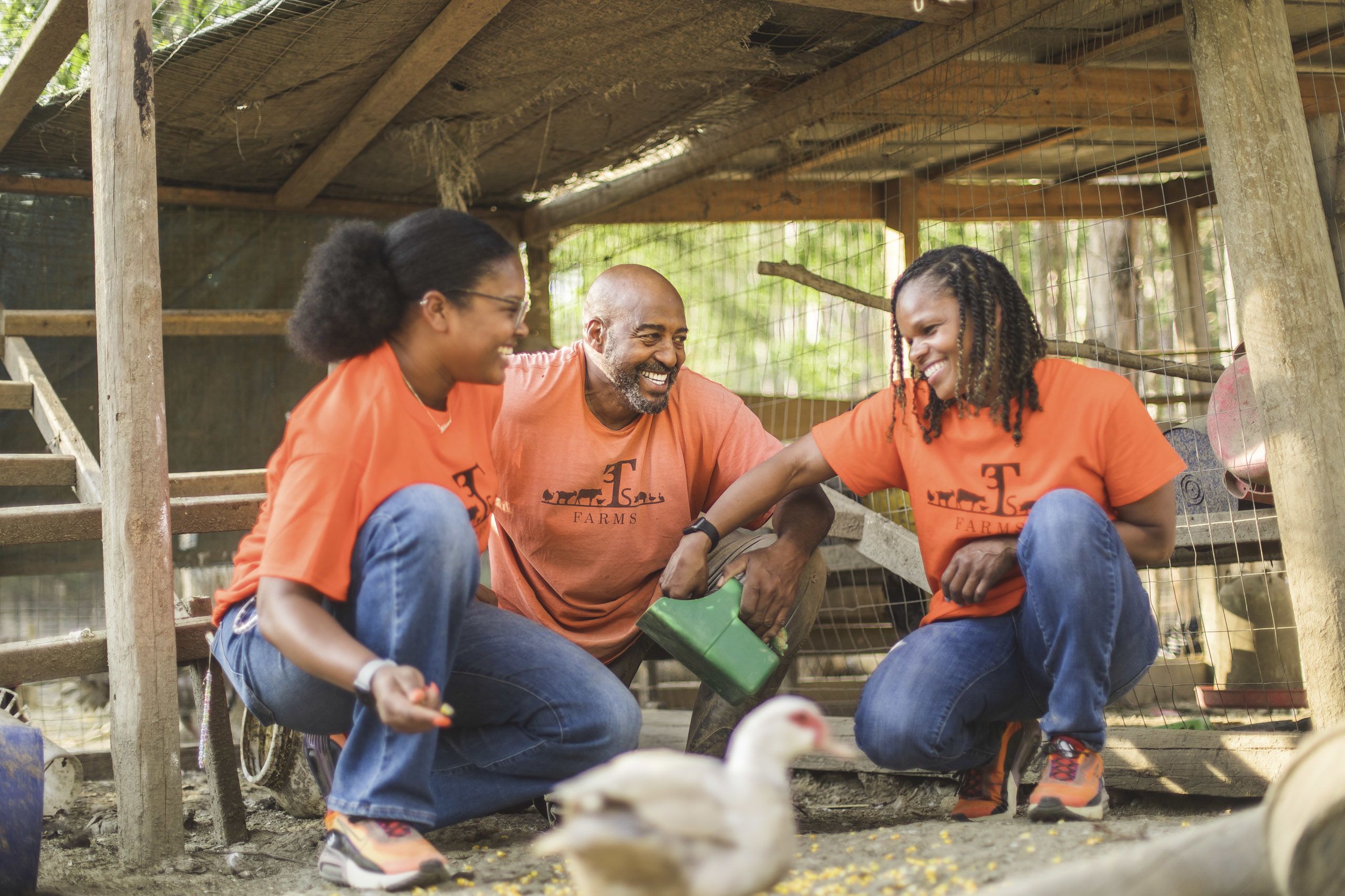 Iteago Felton, his wife, Antheena, and their daughter, Anteaona, feed fowl together on the farm. They are all wearing orange t-shirts that say 3T Farms on them.